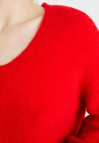 Pull en maille rouge en matériau doux et pelucheux. Il présente un grand col et une coupe décontractée, avec une texture et des coutures visibles.