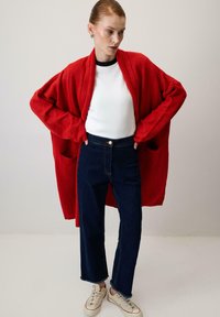 Červený oversize kardigán vyrobený z jemnej látky, kombinovaný s bielou košeľou a tmavomodrými vysokými džínsami. Má dve predné vrecká.