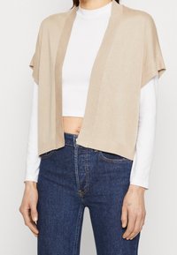 Cardigan a maglia beige con apertura frontale, maniche corte e dettagli a coste. Abbinato a un top bianco e jeans blu scuro.