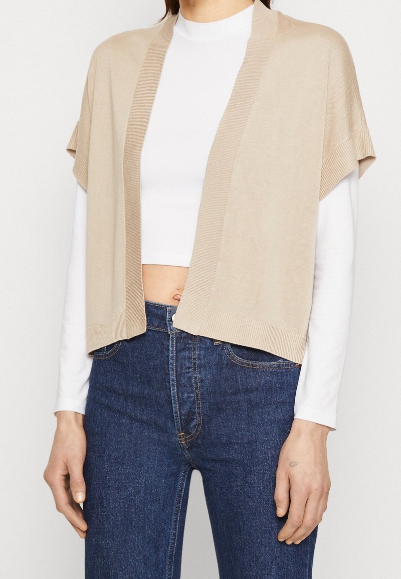 Cardigan a maglia beige con apertura frontale, maniche corte e dettagli a coste. Abbinato a un top bianco e jeans blu scuro.
