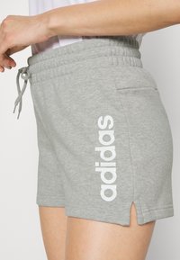 Sive bombažne kratke hlače z vrvico v pasu, stranskimi žepi in razporkom na robu, z velikim belim logotipom "adidas" na strani.