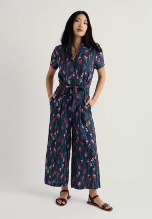 Kobieta stojąca z rękami w kieszeniach, ubrana w niebieski kombinezon w kwiaty z krótkim rękawem oraz brązowe, odkryte sandały na tle jednolitego tła.