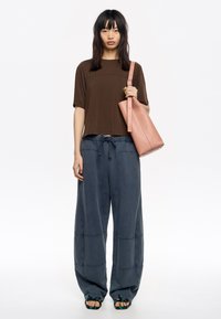 Top marrón de manga corta con tachuelas, combinado con unos pantalones oscuros, sueltos y azules. El modelo lleva un bolso de piel rosa y sandalias negras.