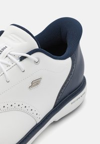 Chaussures de sport blanches et navy avec une tige en synthétique lisse, des détails perforés, un bout rond et un talon foncé contrastant. Présente un logo et un col texturé.