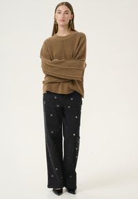 Maglione marrone a coste con maniche lunghe, abbinato a pantaloni neri a gamba larga con occhielli circolari in argento. Scarpe nere a punta completano il look.