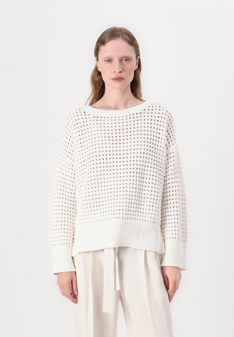 Maglione bianco lavorato a maglia con texture a rete aperta, polsini e orlo a coste, con un logo a patch sul lato inferiore. Abbinato a pantaloni chiari.