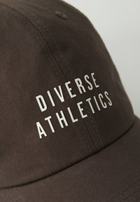 Ciemnobrązowa czapka z materiału, z napisem "DIVERSE ATHLETICS" w białych, pogrubionych literach. Zawiera detale przeszycia oraz guzik na górze.