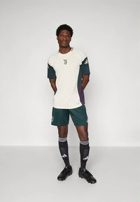 Juventuse jalgpalliriietus beežis ja tumerohelises, millel on trükitud triibud, logodega detailid, lühikesed püksid ja mustad ketsid.