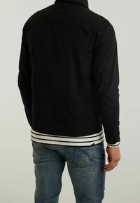 Zwarte denimjas met een kraag, voorzien van een knoopsluiting en een gestreepte witte en zwarte zoom. Draagt over blauwe jeans.
