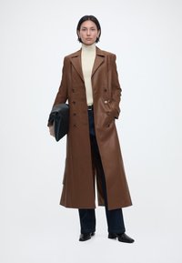 Γυναίκα που φοράει μακρύ καφέ δερμάτινο παλτό, κρεμ πουλόβερ με ζιβάγκο, σκούρο τζιν, μαύρα μπότες, κρατώντας μεγάλη μαύρη τσάντα, στέκεται σε λευκό φόντο.