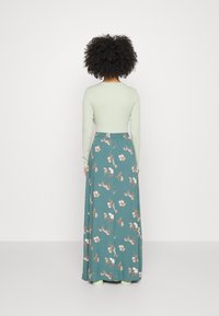 Pitkä kukallinen hame teal-värissä, jossa on vaaleanpunaisia ja valkoisia kukkakuoseja, yhdistettynä vaaleanvihreään pitkähihaiseen puseroon, tiukalla vyötäisellä ja kelluvalla muotoilulla.