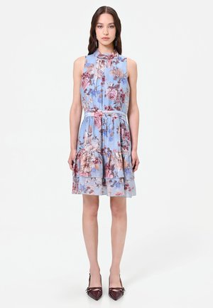Femme portant une robe légère sans manches bleu clair à motif floral avec un ourlet volanté et une ceinture assortie, associée à des chaussures pointues rouge foncé, debout sur fond blanc.