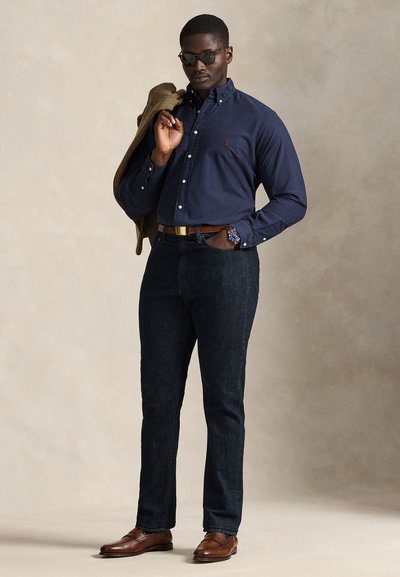 Vyras dėvintis tamsiai mėlyną marškinių su sagutėmis, tamsias džinsines kelnes, rudą diržą ir batus, saulės akinius, laiko rudą švarką ant peties.