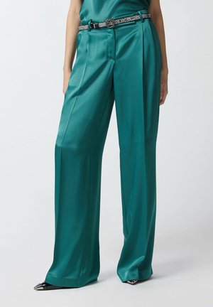 Pantalons en satin teal à jambes larges avec plis frontaux, accompagnés d'une ceinture noire ornée de strass et de chaussures noires à bouts pointus.