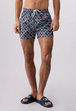 Uomo che indossa pantaloncini da bagno blu navy con motivo e coulisse bianca, e sandali scivolati blu navy con logo bianco su sfondo neutro.