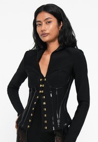 Γυναίκα με μαύρα κύματα μαλλιά, φορώντας ένα σκουρόχρωμο προσαρμοσμένο σακάκι με χρυσά κούμπωματα και γυαλιστερές κατακόρυφες ρίγες, ποζάροντας μπροστά σε άσπρο φόντο.