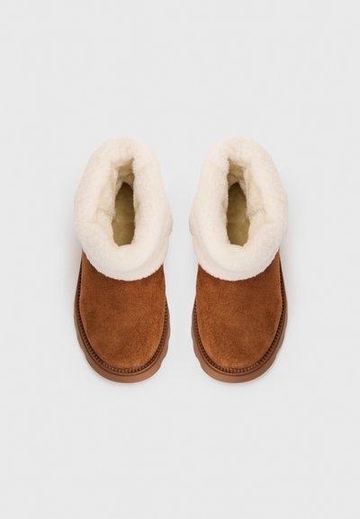 Chaussons en nubuck marron avec une épaisse doublure en fausse fourrure blanche et une semelle antidérapante, disposés côte à côte sur un fond blanc uni.