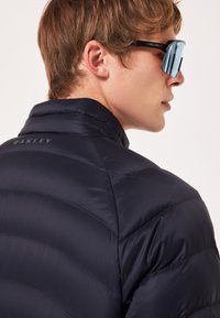 Marineblå pufferjakke med kviltet design, høj Krave og "Oakley" logo på ryggen. Båret med oversized solbriller. Glat tekstur.