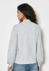 Grau gestrickter Pullover mit hohem Kragen, Ballonärmeln und gerippten Bündchen. Hergestellt aus einem weichen, strukturierten Stoff. Rückansicht gezeigt.