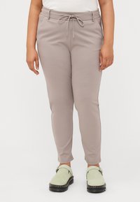 Helle beige Hose mit einem Kordelzugbund, kombiniert mit grünen Slippern, getragen von einer Person, die vor einem weißen Hintergrund steht.