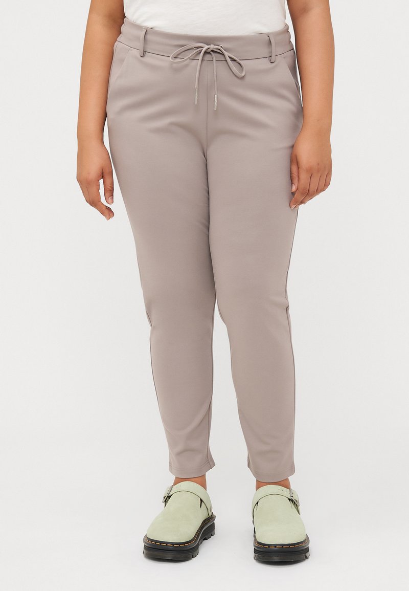 Helle beige Hose mit einem Kordelzugbund, kombiniert mit grünen Slippern, getragen von einer Person, die vor einem weißen Hintergrund steht.