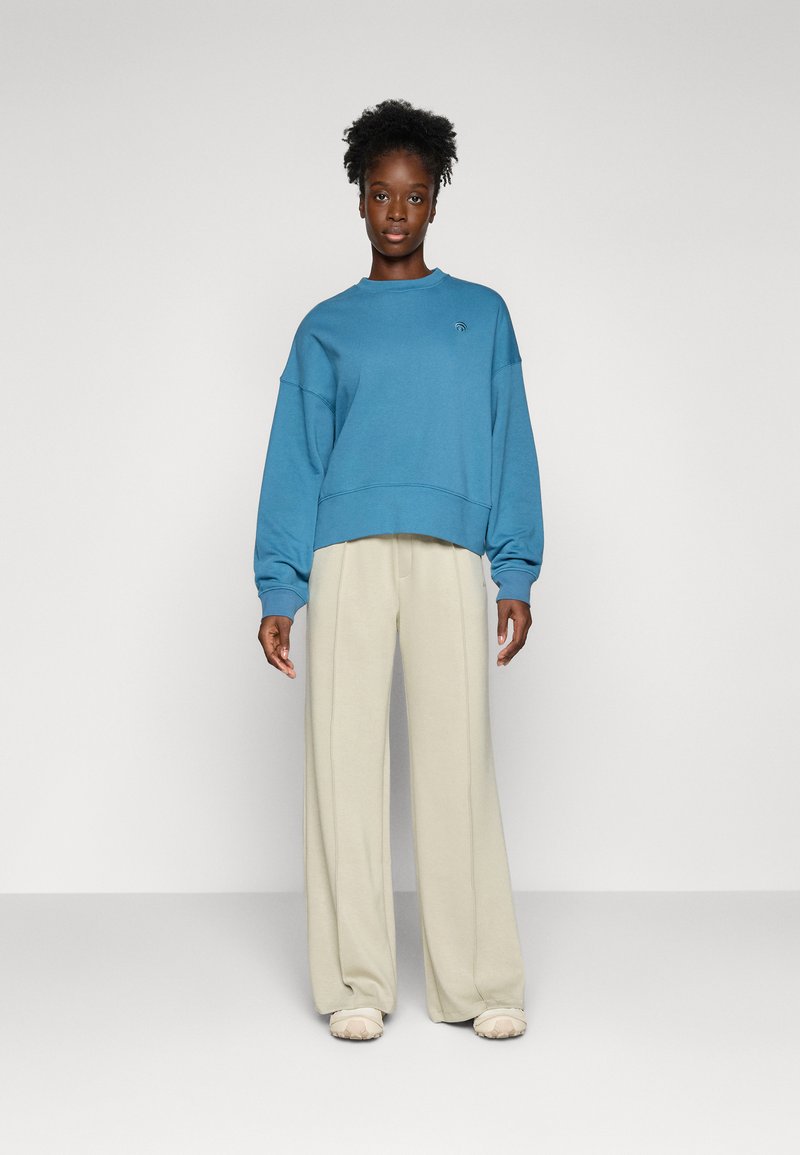 Felpa azzurro chiaro con vestibilità rilassata e collo rotondo, abbinata a pantaloni a gamba larga color crema, presentata su uno sfondo bianco semplice.