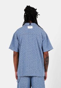 Blauwe shirt met korte mouwen en een gemusterde textuur. Heeft een label aan de achterkant. Gecombineerd met bijpassende shorts in een vergelijkbaar patroon.