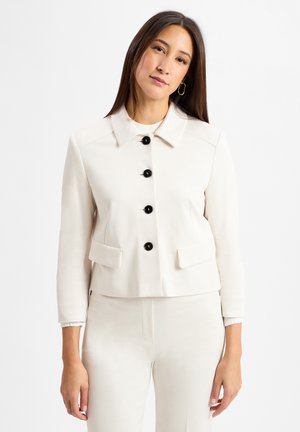 Frau mit langen braunen Haaren, die ein weißes Blazer mit schwarzen Knöpfen und passende weiße Hose trägt, steht vor einem weißen Hintergrund.