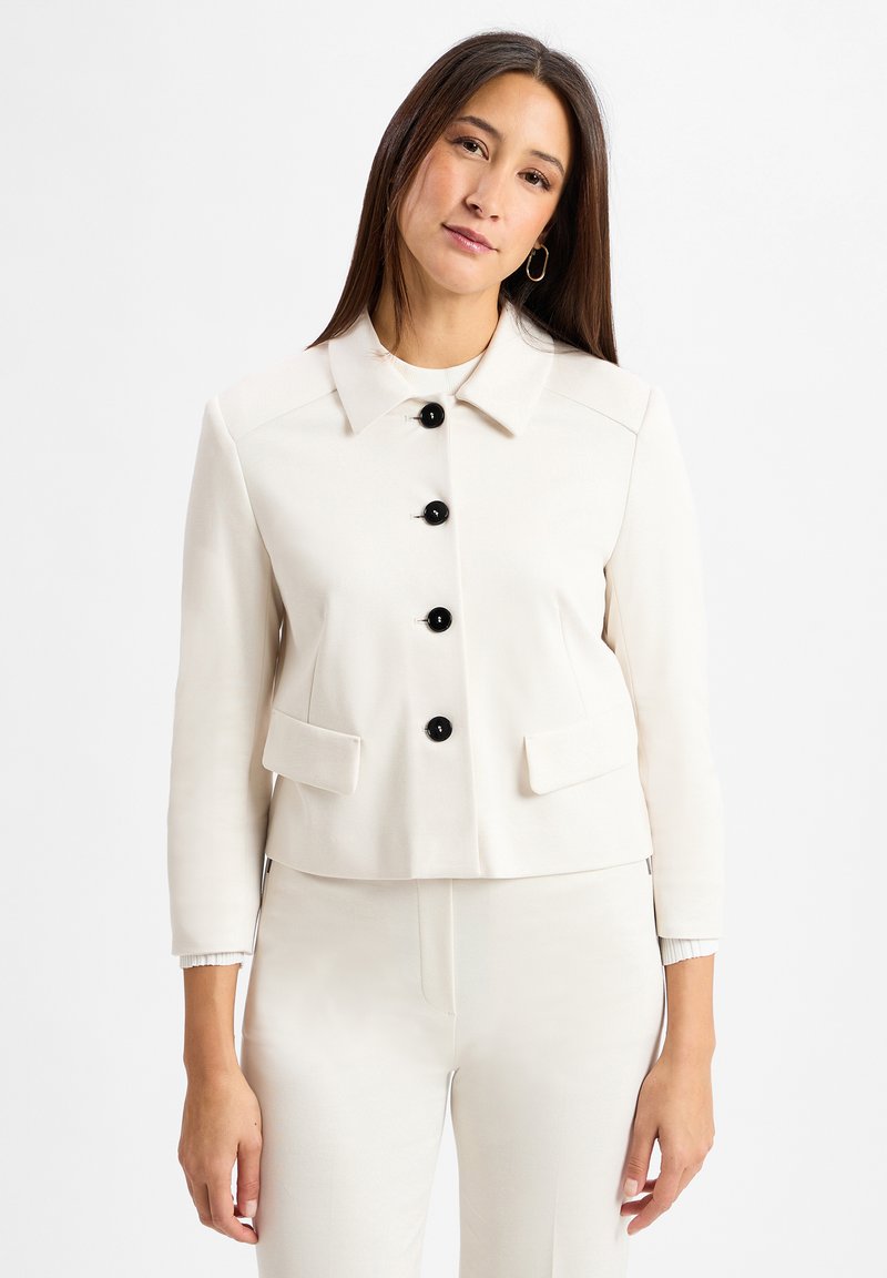 Frau mit langen braunen Haaren, die ein weißes Blazer mit schwarzen Knöpfen und passende weiße Hose trägt, steht vor einem weißen Hintergrund.