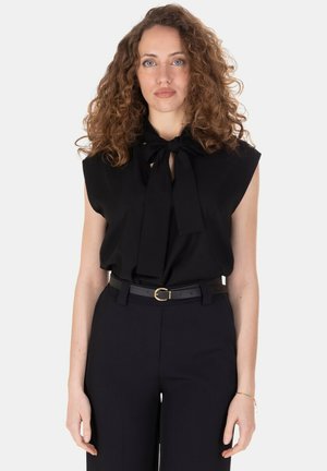 Donna con capelli ricci che indossa una camicetta nera senza maniche con fiocco e pantaloni neri con cintura, in piedi su sfondo bianco.