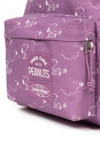 Mochila de tela morada con personajes de Snoopy en blanco y patrones florales, con un bolsillo delantero que muestra etiquetas de marca y un cierre de cordón.