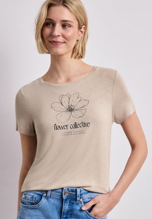 Blondynka w beżowym t-shircie z czarnym szkicem kwiatu i napisem "flower collective", w połączeniu z niebieskimi dżinsami, pozująca na jednolitym tle.