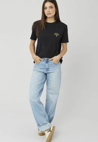 Femme aux longs cheveux bruns portant un T-shirt noir "Start Creating", un jean large bleu clair et des baskets blanches, debout les mains dans les poches.