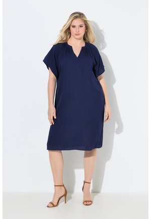 Vrouw met blond haar die een marineblauwe jurk tot op de knieën draagt met korte mouwen en bruine sandalen met hoge hakken, staand tegen een witte muur.