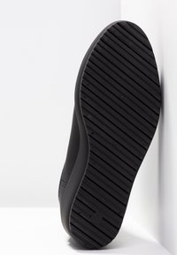 Semelle d'une chaussure noire avec des rainures diagonales, reposant verticalement contre un mur d'angle blanc.