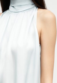 Haut sans manches en satin bleu clair avec un col montant froncé porté par une personne aux longs cheveux foncés, dévoilant l'épaule et le haut de la poitrine.