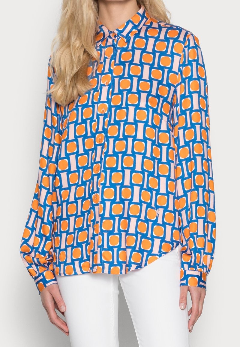 Femme portant une chemise à manches longues boutonnée avec un motif géométrique bleu, orange et rose, associée à un pantalon blanc.