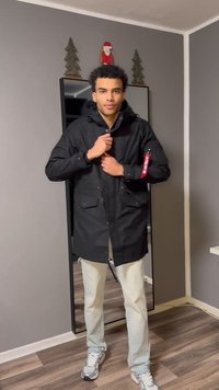 Musta vedenpitävä parkatakki, jossa on huppu, kaksi etutaskua ja punainen yksityiskohta hihassa. Päällä vaaleat housut ja lenkkarit.