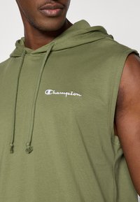 Αμάνικο φούτερ με κουκούλα σε ελιά πράσινο, κατασκευασμένο από βαμβάκι, με ρυθμιζόμενη κουκούλα και  κεντητό λογότυπο "Champion" σε λευκό στο στήθος.