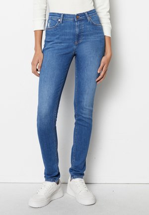 Person iført mellemhøjt, tætsiddende blå jeans kombineret med hvide sneakers og en hvid langærmet top mod en ensfarvet baggrund.