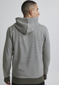 Grijze hoodie van een textiel met structuur, voorzien van een trekkoord in de capuchon en groene ribgebreide manchetten en onderrand. De achterkant heeft een simpel, strak ontwerp.