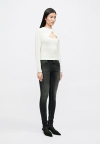 Λευκή μπλούζα μακρυμάνικη με μοναδικό σχέδιο κοπής, συνδυασμένη με μαύρα τζιν στενής γραμμής και μαύρες παπούτσια με尖尖 toe. Λείος υφή συνολικά.