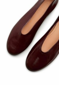 Burgundowe skórzane baleriny o gładkim wykończeniu, z zamkniętymi palcami i wycięciami po bokach, odsłaniającymi jasnobrązowe wnętrze.