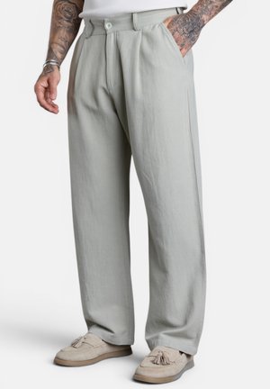 Man wearing light gray wide-leg trousers and beige tassel loafers, with tattooed arms and one hand in pocket.