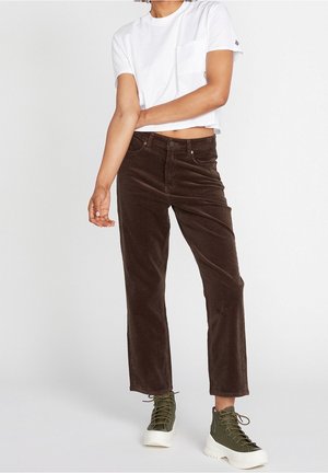 Personne portant un t-shirt court blanc, un pantalon en velours côtelé marron et des baskets compensées vert olive, debout devant un fond uni.