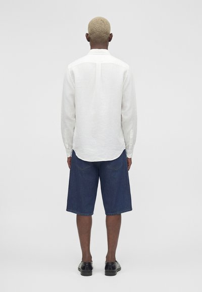 Άνδρας με κοντά ξανθά μαλλιά που φοράει λευκό πουκάμισο με μακριά μανίκια, σκούρες μπλε τζιν σορτς και μαύρα επίσημα παπούτσια, στέκεται γυρισμένος προς τα πίσω.