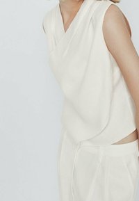 Crème-kleurige mouwloze blouse met een gewikkeld ontwerp, gemaakt van een gladde stof. Beschikt over een hoge halslijn en een ontspannen pasvorm. Gecombineerd met vergelijkbare broeken.