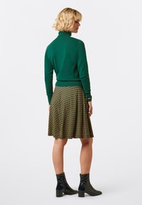 Donna con capelli biondi corti che indossa un dolcevita verde, una gonna a pieghe a lunghezza ginocchio con pattern e stivali neri in vernice fino alla caviglia, in piedi su uno sfondo bianco.