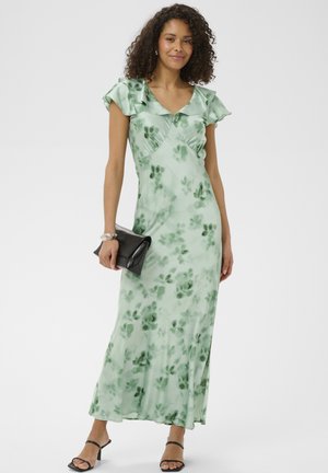 Vrouw die een lichtgroene maxi-jurk met bloemenprint en gerufflede mouwen draagt, een zwarte clutch vasthoudt en zwarte sandalen met hakken aan heeft.