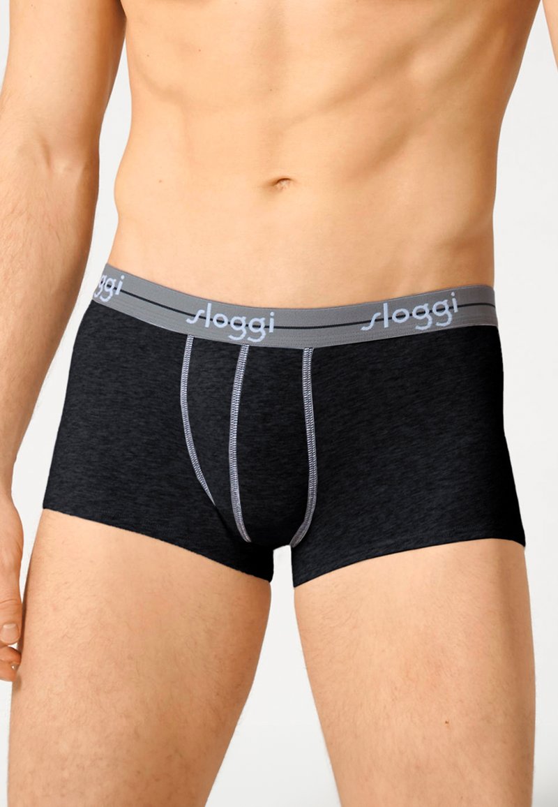 Torso maschile che indossa boxer neri con una fascia grigia con il marchio "sloggi" su uno sfondo chiaro e semplice.
